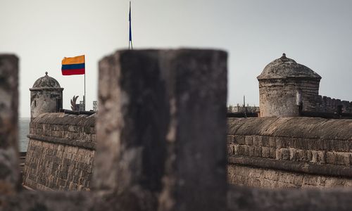 A historic stone fortress near the sea, featuring a small watchtower and a waving Colombian flag. Blurred stone structures frame the scene, adding depth and focus.